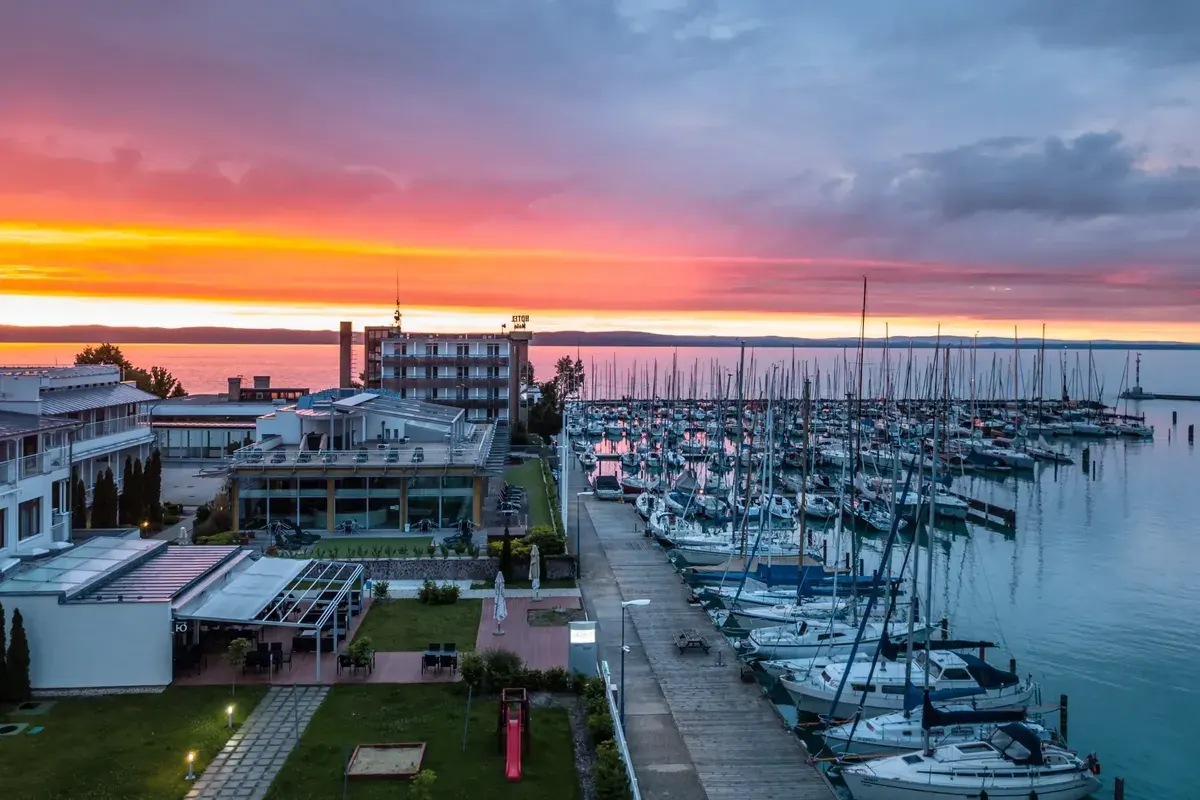Esti légi felvétel a Hotel Yacht Siófokról rózsaszín-narancssárga égbolttal, ahol a vitorláskikötő és a Balaton látható naplementében
