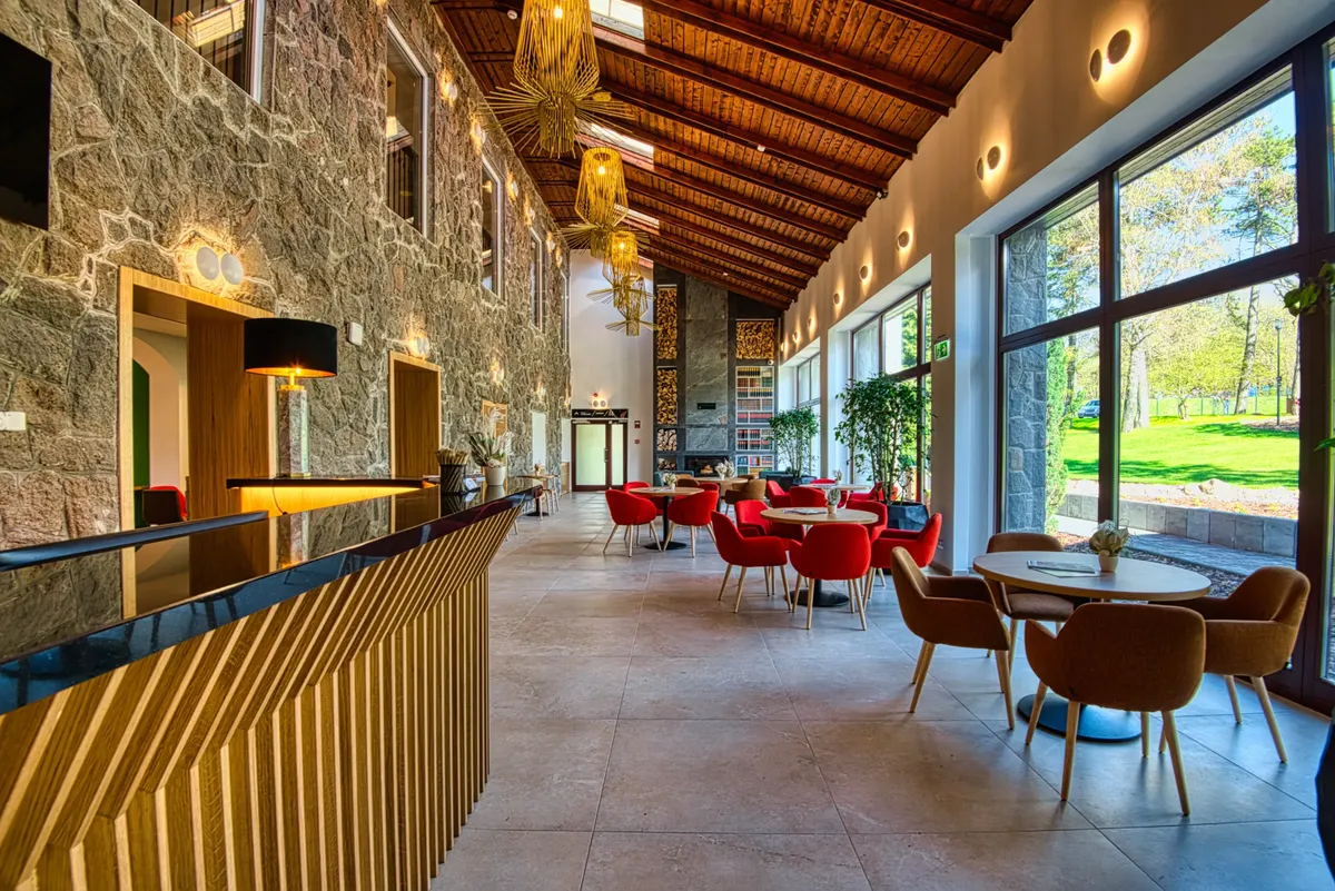 A Cardoner Hotel Dobogókő lobby-ja, ahol terméskő burkolat, fatüzelésű kandalló, fa gerendás mennyezet és designer csillár látható panorámaablakokkal az erdőre