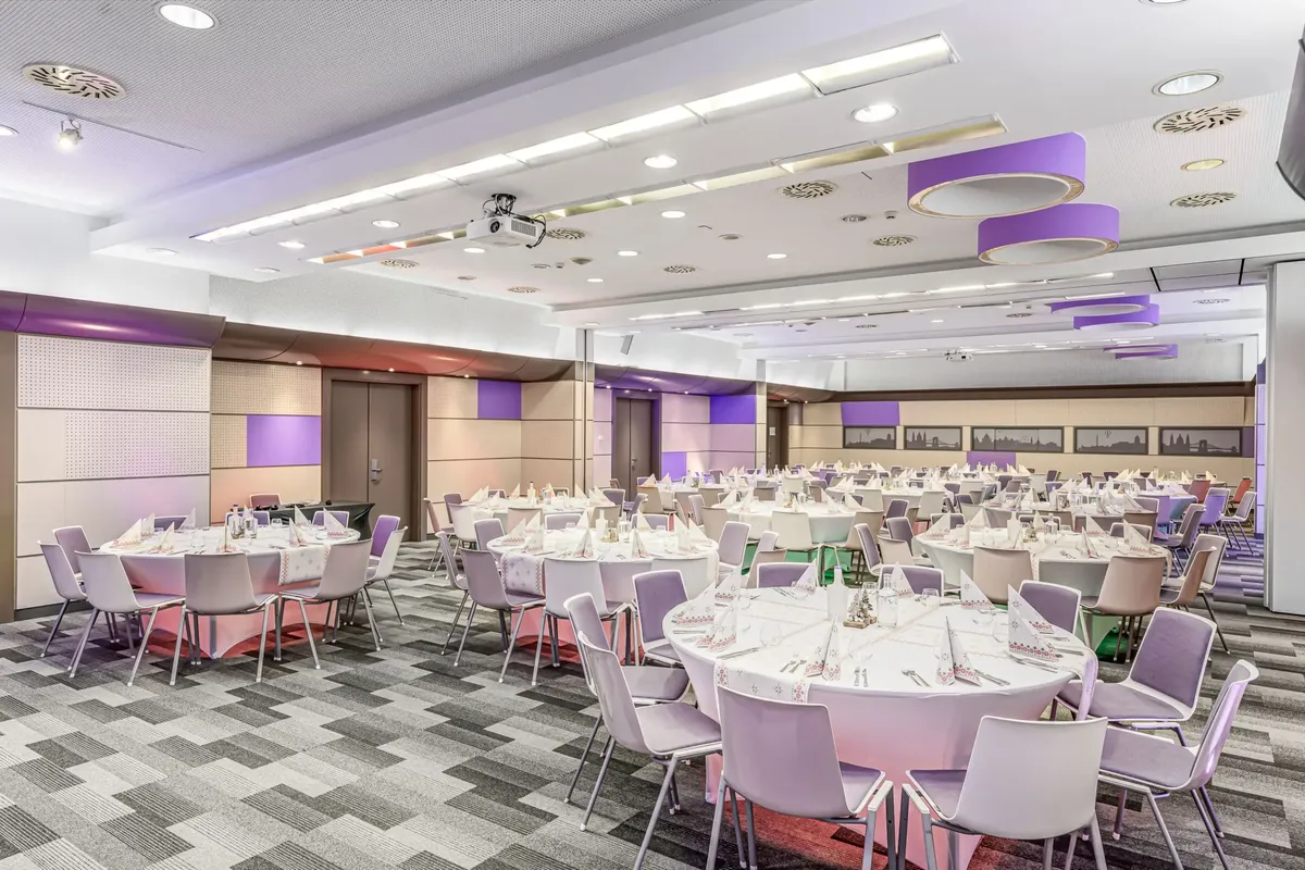A Mercure Budapest Castle Hill Mátyás I-II. egyesített terme körasztalos elrendezéssel, ahol 130 fő vesz részt gálavacsorán lila LED világítással és beépített projektorral